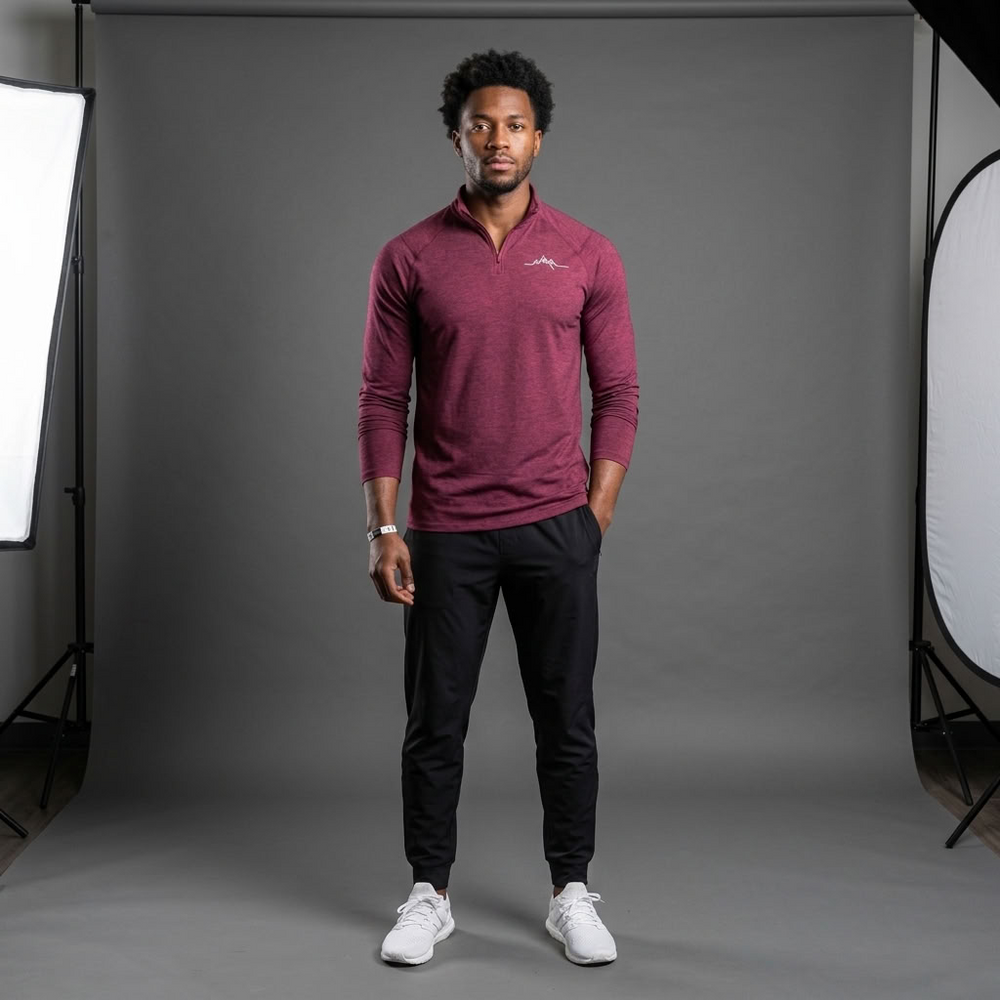 A man stands in a studio wearing a heathered burgundy long-sleeve quarter-zip pullover with a small white logo on the left chest, paired with black joggers and white sneakers.